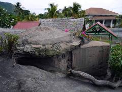 The image shows a weathered concrete bunker surrounded by vegetation, with buildings and palm trees in the background, likely part of the Rabaul WWII Memorial.