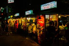 A vibrant night scene unfolds at a bustling Japanese food stall, illuminated by glowing signs and lanterns, where patrons gather to enjoy ramen and other culinary delights.