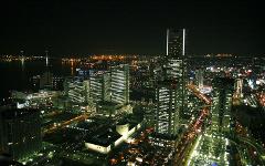 This is an aerial shot of a city at night, featuring several lit-up skyscrapers and a bay with a bridge in the background.