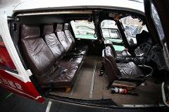 Inside a helicopter cabin: brown leather seats with seatbelts face the cockpit with instruments and controls. Another helicopter is seen outside.