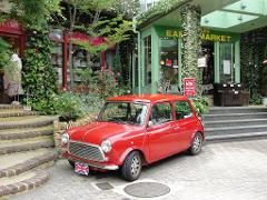 A vintage red Mini Cooper is parked on a brick-paved street in front of a building with lush greenery, floral decorations, and shop signs in both English and Japanese.