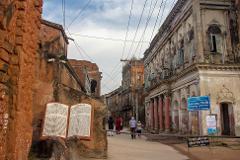 An eye-level shot of the street leading into Panan City in Bangladesh, with a sign explaining the history of the place and another sign mandating the use of masks.