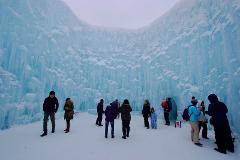 A group of people are gathered at the base of a massive, blue ice formation with icicles.
