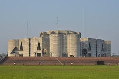 The Bangladesh National Parliament House stands prominently with its geometric facade and cylindrical structures, set against a clear sky and vast green field.