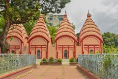 Four quaint, peach-colored temples with red-striped conical roofs stand behind a decorative fence, set against a backdrop of lush trees and a modern building.