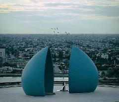 The image shows the Al-Shaheed Monument in Baghdad, Iraq, featuring two split turquoise domes with a distant cityscape and birds in flight.