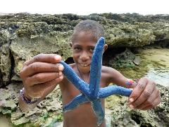 A young boy stands smiling, holding a blue starfish in front of a rocky shoreline.