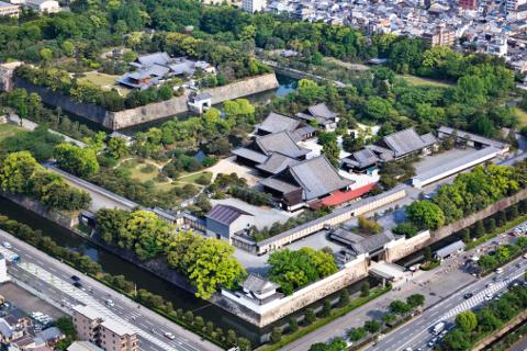 Flygfoto över ett traditionellt japanskt slott omgivet av en vallgrav, murar och frodig grönska, med stadsbyggnader synliga i bakgrunden.