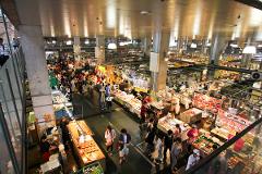 A busy marketplace filled with vendors and shoppers, viewed from above, with bright overhead lighting.