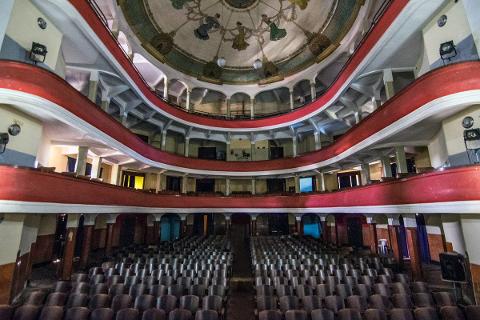 A grand auditorium with rows of empty seats and a red-and-white balcony under an ornate, dome-shaped ceiling featuring figures.