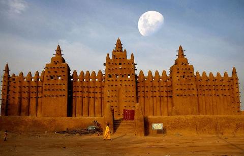 The Great Mosque of Djenné stands tall under a bright moon as a figure in yellow walks toward its mud brick facade.