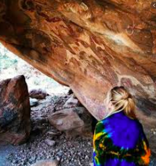 A woman in a colorful shawl stands under a rock overhang with ancient rock art.