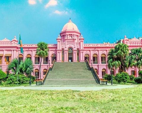 A grand pink palace with a dome and a wide staircase stands under a bright blue sky, surrounded by lush green grass and trees.