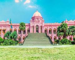 A grand pink palace with a dome and a wide staircase stands under a bright blue sky, surrounded by lush green grass and trees.