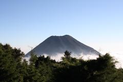 A large, gray volcano rises above the cloud line and green trees against a clear blue sky.
