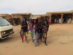 A group of six people, including one child, are walking in a desert-like setting with buildings and a van in the background.