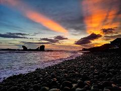 A stunning sunset over a rocky beach with dramatic clouds and colorful light streaking across the sky.