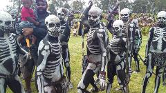 A group of people, painted in black and white skeleton designs, stand on a grassy field with a man holding a child in the background.