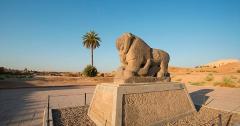 An ancient stone lion statue stands prominently in a desert landscape, with a palm tree nearby and buildings in the distance under a clear blue sky.