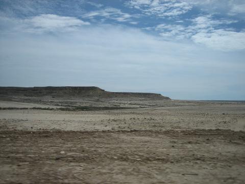 A vast, arid landscape stretches out under a partly cloudy sky, with a low, flat-topped mountain range in the distance.