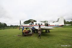 A small white and red P2-TNT airplane sits on a grassy field, with several people loading and unloading cargo.