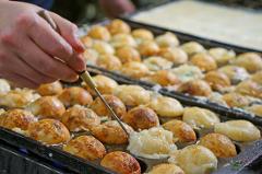 A person uses a skewer to prepare takoyaki, a Japanese snack of battered octopus balls, in a specialized pan with round molds.