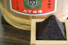 Step Inside a 400-Year-Old Miso Warehouse — The Hatcho Miso Experience in Okazaki, Aichi A pyramid of dark miso sits in a wooden frame in front of a blurred, out-of-focus background showing a large wooden barrel with a red label featuring Japanese text.