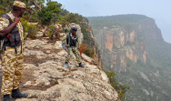 Two armed security guards in camouflage uniforms stand on a rocky cliff overlooking a hazy, mountainous landscape.