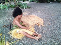 A woman with an afro sits on a rocky ground wearing a leaf skirt and tying light-colored grass in Alotau, Milne Bay.
