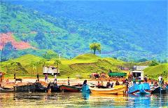 A vibrant landscape featuring a riverbank with colorful boats, a lush green hill, and people engaged in daily activities.