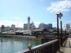 A cityscape featuring traditional Japanese buildings along a waterway, with modern structures and a communication tower in the background, under a bright blue sky.