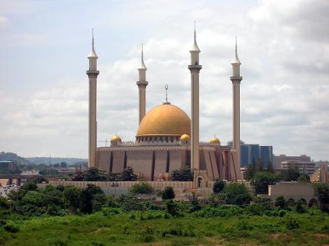 A grand mosque with golden dome and tall minarets rises above lush greenery under a cloudy sky.