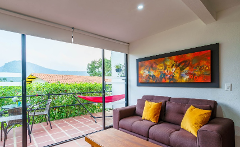 A living room with a brown couch with yellow pillows, an abstract painting, and a sliding glass door that leads to a balcony with a table, chairs, and a red hammock overlooking a green landscape.