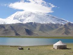 A scenic view of a lake with a yurt on the shore, set against a backdrop of majestic snow-capped mountains under a blue sky.