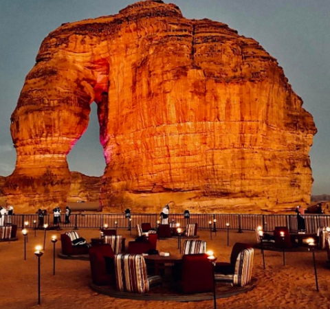 An outdoor lounge with comfortable chairs and torches sits at the base of a large, striking rock formation with an archway.