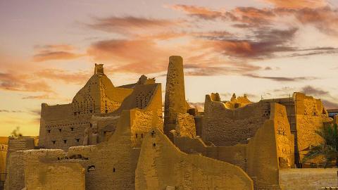 The image shows the historic mud-brick architecture of At-Turaif, a UNESCO World Heritage Site in Saudi Arabia, set against a dramatic sunset sky.