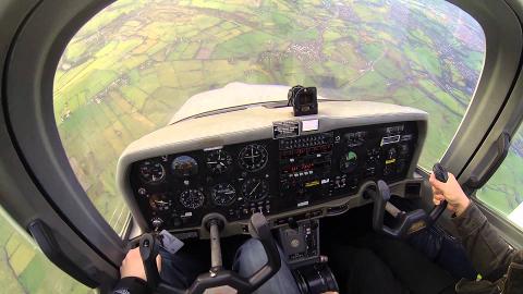 An eye-level shot of a person's view from inside a small aircraft mid-flight, showing the pilot holding the plane's control. The aircraft is tilted, revealing a green landscape below through the window.