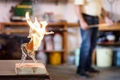 A blurred image shows a glass sculpture of a leaping deer with flame-like accents on a wooden base, with a glass artisan in the background working.