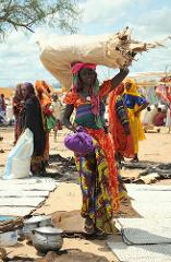 A woman carrying a large sack on her head stands in a busy marketplace with other people and colorful clothing.