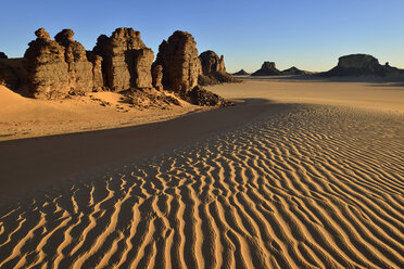 A desert landscape showcases wind-swept sand dunes and rocky formations under a clear, blue sky in a scene evoking arid beauty and isolation.