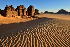 A desert landscape showcases wind-swept sand dunes and rocky formations under a clear, blue sky in a scene evoking arid beauty and isolation.