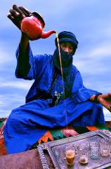 A man in a blue robe and head wrap pours tea from a red teapot into glasses on a silver tray, likely in a desert setting.