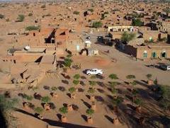 Here's a one-sentence description of the image:An aerial view of a remote desert village with mud-brick buildings, scattered vegetation, and a white vehicle in a central area.