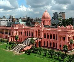 A pink palace with a large staircase is set against a backdrop of green lawns, trees, and a cloudy sky, surrounded by city buildings.
