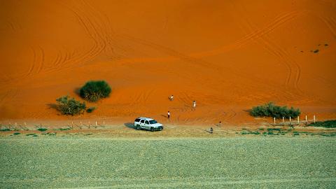 Aerial shot of a white SUV parked on the border of a green field and a large orange sand dune with scattered plants and several people walking on it.