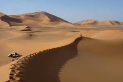 A vast desert landscape with rolling sand dunes, a white truck parked at the base, and a lone person sitting atop a distant dune crest.