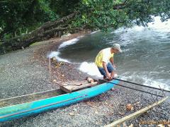 A man in a yellow shirt and cap launches a small, bright blue outrigger canoe into the water from a rocky beach lined with trees.