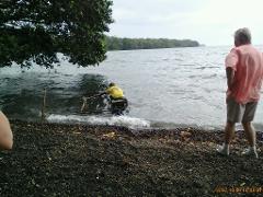A man in a yellow shirt wades into the water pulling a structure as another man in a pink shirt observes from the rocky shore.