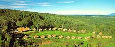 Aerial view of Ambua Lodge, a collection of thatched-roof bungalows nestled in a lush, green valley surrounded by dense rainforest.