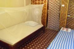 A room with two beds; one with a mosquito net and a brown frame, and the other with a blue and white checked pattern.